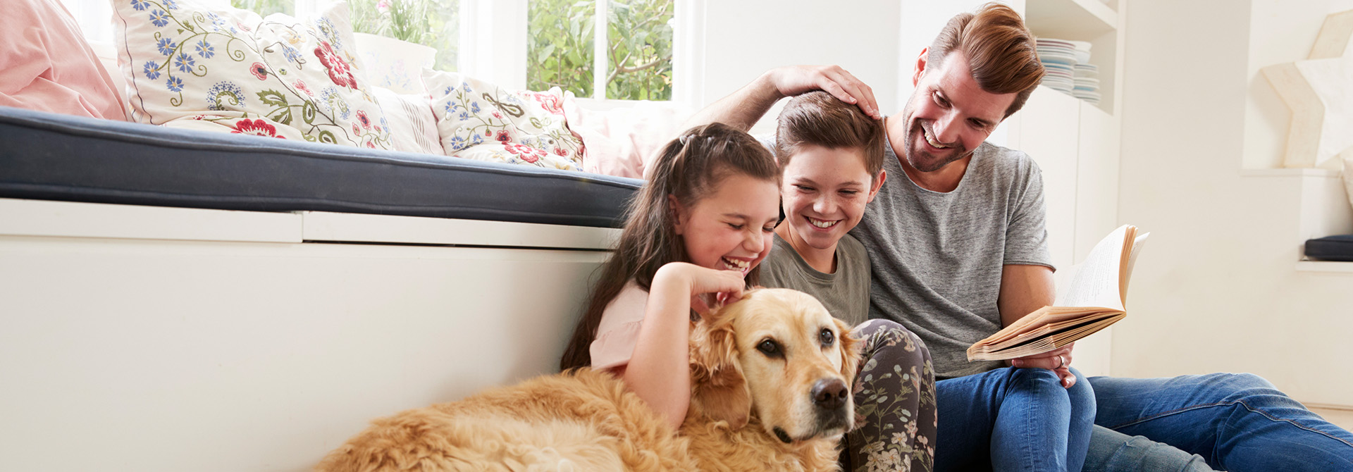 Zwei Kinder sitzen auf dem Boden neben ihrem Papa, der ein Buch in der Hand hält und einem ruhigen Golden Retriever in einem hellen Wohnzimmer